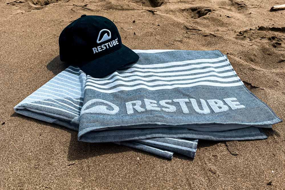 Handtuch liegt am Strand gefaltet und auf der Ecke liegt eine Kappe auf der RESTUBE steht