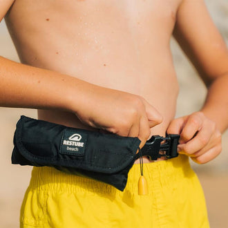 Boy in yellow swimming shorts puts his Restube beach swimming buoy on belt around waist